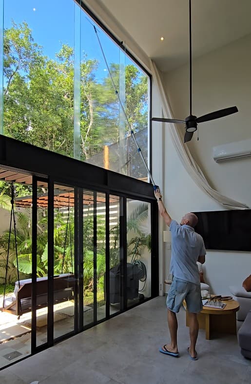 Qtana cleaner using an extension pole to clean floor-to-ceiling windows in a jungle-view living room